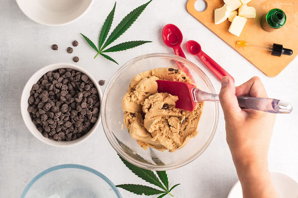 Hand stirring cookie dough with chocolate chips and marijuana leaves around bowl