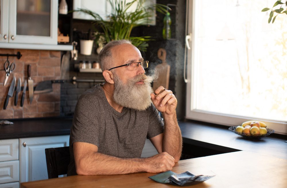 A senior man smokes cannabis sitting at his kitchen table